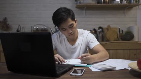 fast forward japanese male sitting behind laptop is writing notes while calculating living cost with a calculator at dining table in the apartment at night.