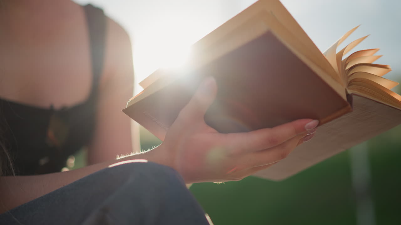 los rayos del sol se reflejan en el libro abierto sostenido por una mujer sentada al aire libre con las piernas cruzadas, mientras el viento sopla, las páginas giran suavemente, mientras que el fondo y la mujer están ligeramente borrosos