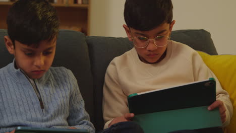 two young boys sitting on sofa at home playing games or streaming onto digital tablets 4