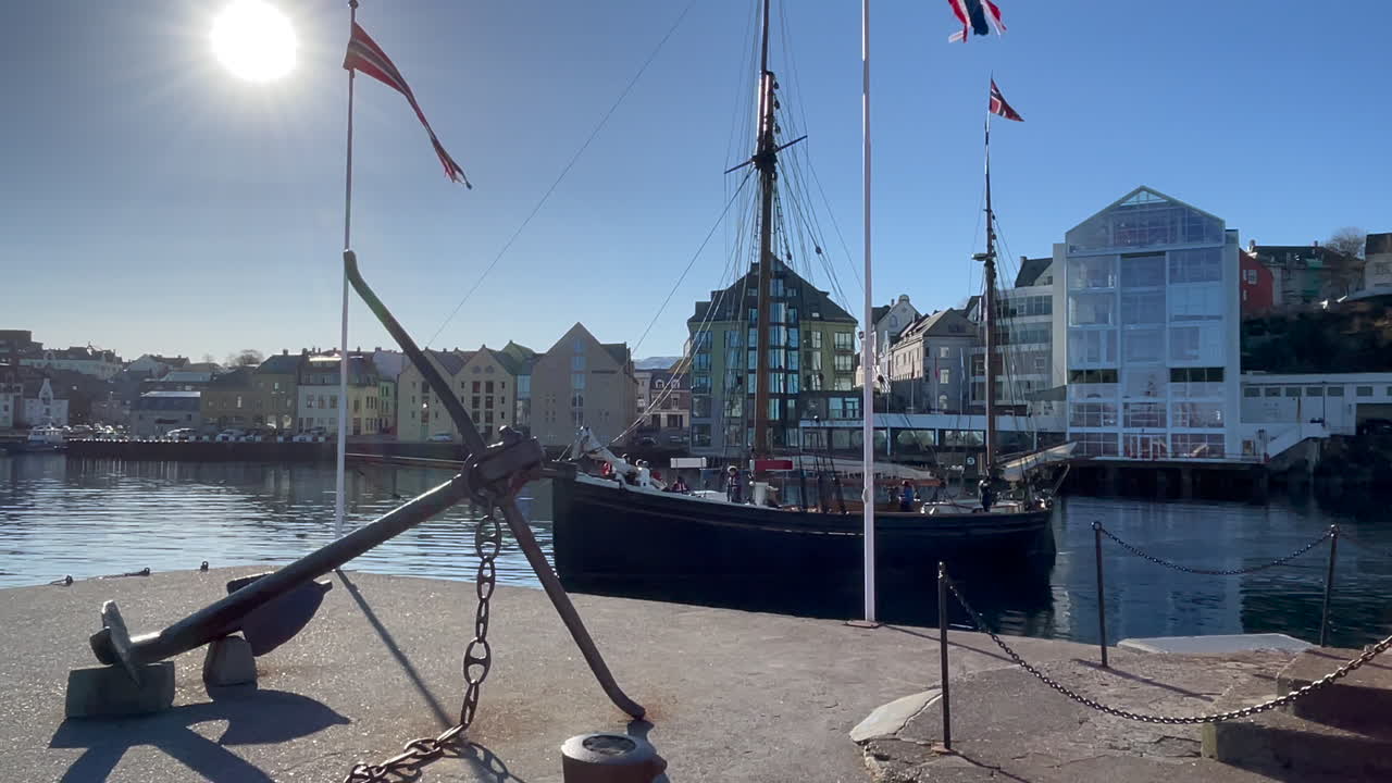 viejo barco de madera que sale del puerto de ålesund, pasado el ancla y el faro de molja, el hotel y la academia thon, sol brillante, panorámica de derecha a izquierda