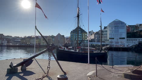 Viejo-Barco-De-Madera-Que-Sale-Del-Puerto-De-Ålesund,-Pasa-El-Ancla-Y-El-Faro-De-Molja,-El-Hotel-Y-La-Academia-Thon,-Sol-Brillante,-Panorámica-De-Derecha-A-Izquierda