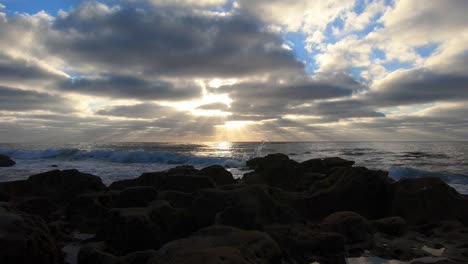 establecimiento de una toma de rayos solares que alcanzan su punto máximo a través de las nubes sobre el océano en un día nublado con pequeñas olas rodando hacia la orilla con rocas en primer plano y parches de cielo azul