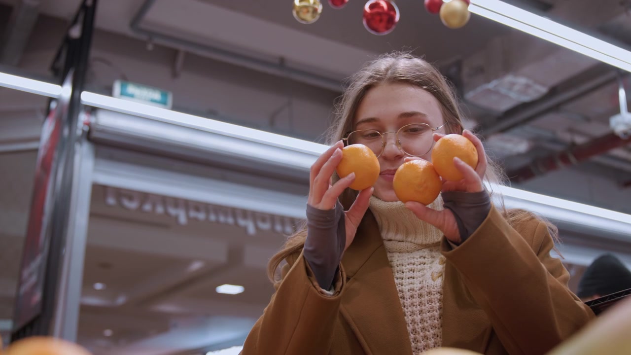 Donna bianca che confronta arance al mercato. Luminosa esposizione di agrumi con ornamenti festivi in alto, mani che ispezionano la consistenza, cliente sorridente e giocosa che valuta la scelta tra mandarino e mango, colorato.