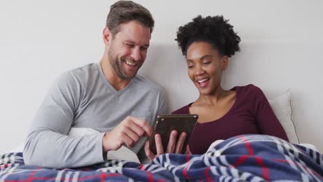 video of happy diverse couple using tablet in bed and talking