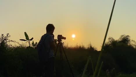 Silhouette-Eines-Fotografen,-Der-Den-Sonnenuntergang-Mit-Einer-DSLR-Und-Einem-Stativ-Auf-Einer-Wiese-Vor-Der-Untergehenden-Sonne-Fotografiert