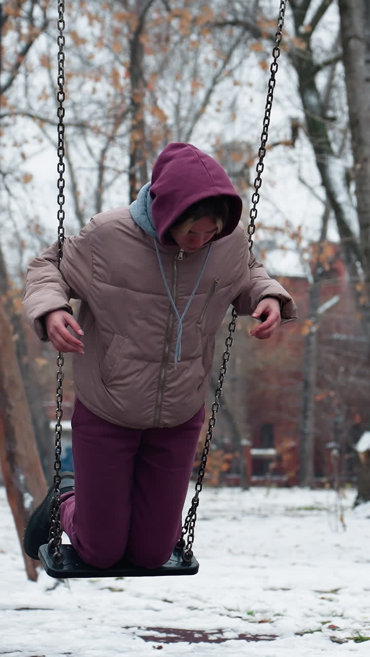 dama vestida cálidamente para el invierno en el columpio en el parque sosteniendo cadenas, con pantalones de color marrón y chaqueta de invierno, con nieve en el suelo y árboles en el fondo