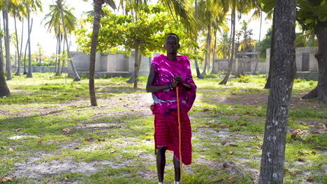 Aldeano-Africano-Vestido-De-Rosa-Y-Con-Palo-Parado-En-Un-Palmeral