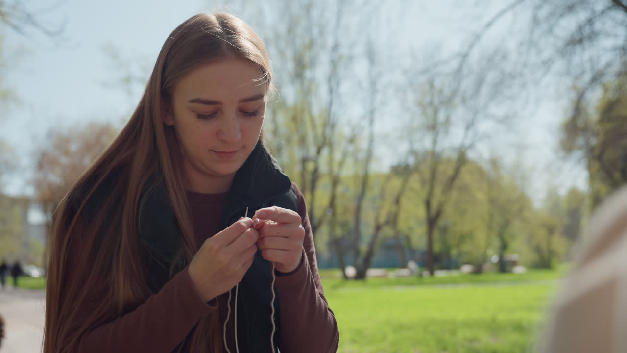 Молодой кавказский инфлюенсер тестирует наушники, сидя на скамейке, игриво улыбается, поправляет шнуры, на фоне парк, весенний свет, повседневный свитер, смотрит в камеру, в творческом настроении, случайный портрет