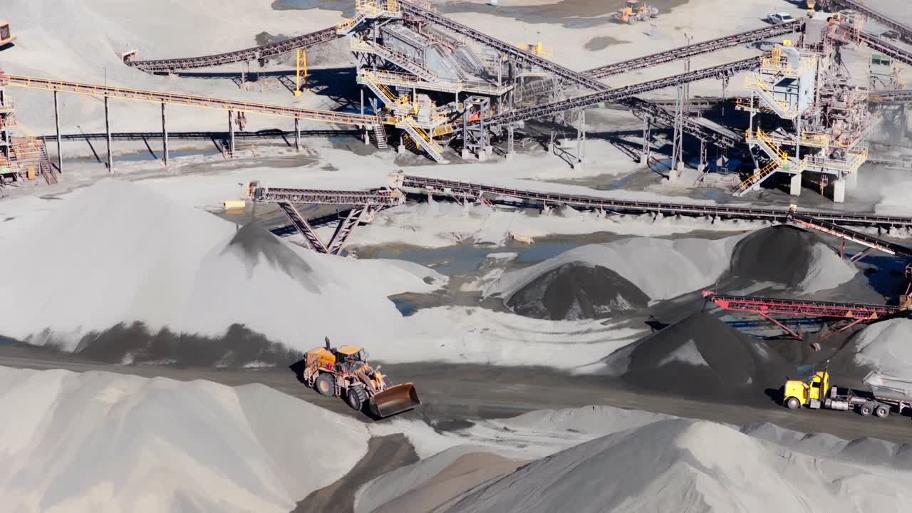 Wheel loader driving across quarry with aggregate processing plant conveyor belt system, active worksite, aerial