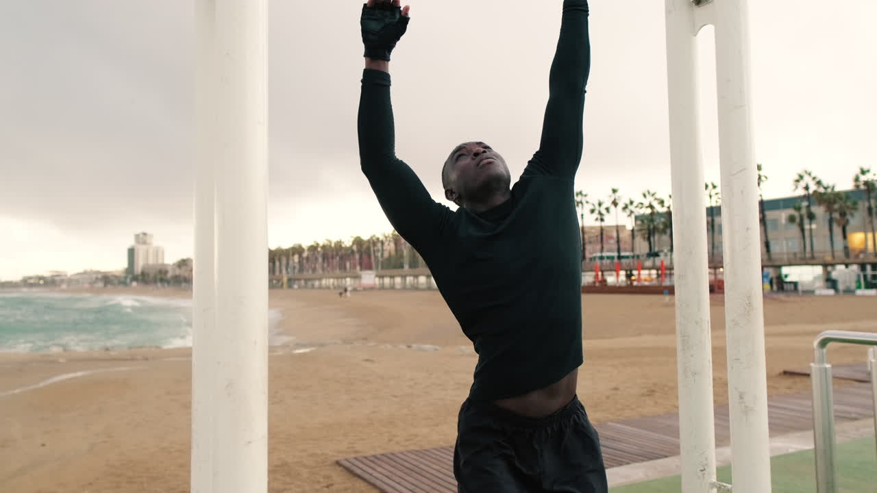 Young Black Man Stretching and Hanging on Pull-Up Bar at the Beach