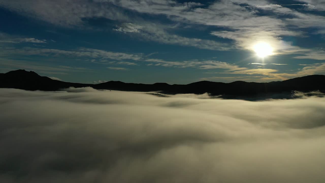 vuelo de avión no tripulado sobre un mar de nubes con el sol en frente visualizando las montañas que parecen oscuras de la luz en un giro a la derecha con un cielo azul en un amanecer en invierno en ávila, españa