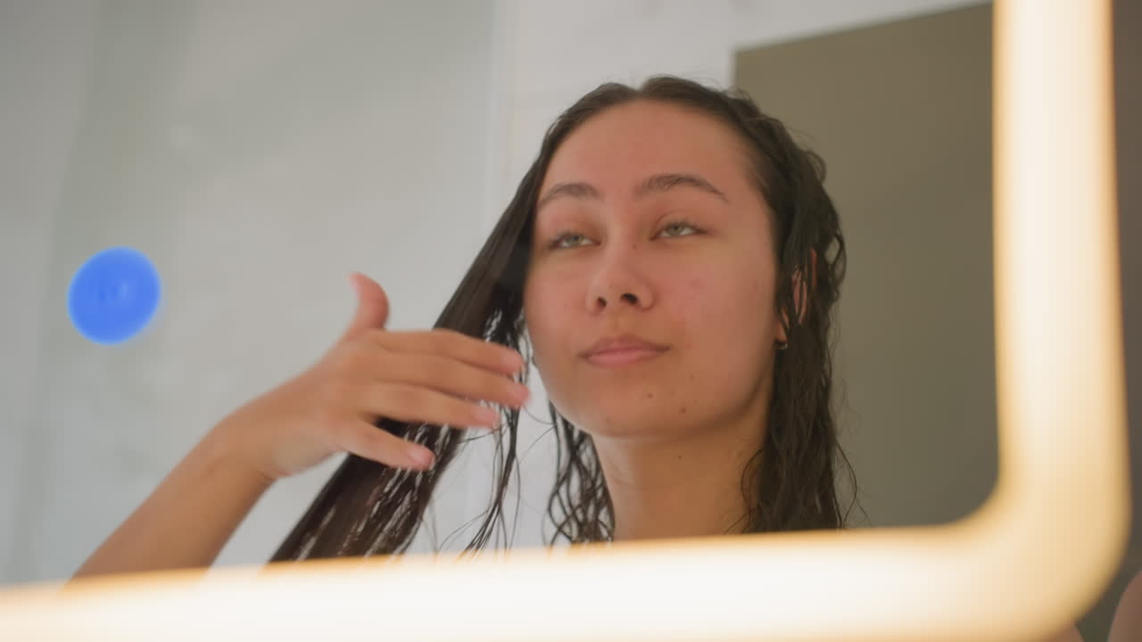Woman grooming herself. Woman beautifying in bathroom mirror. Selfcare routine in cozy hotel bathroom. Relaxing selfcare session involving hair brushing and mirror gaze in warm light
