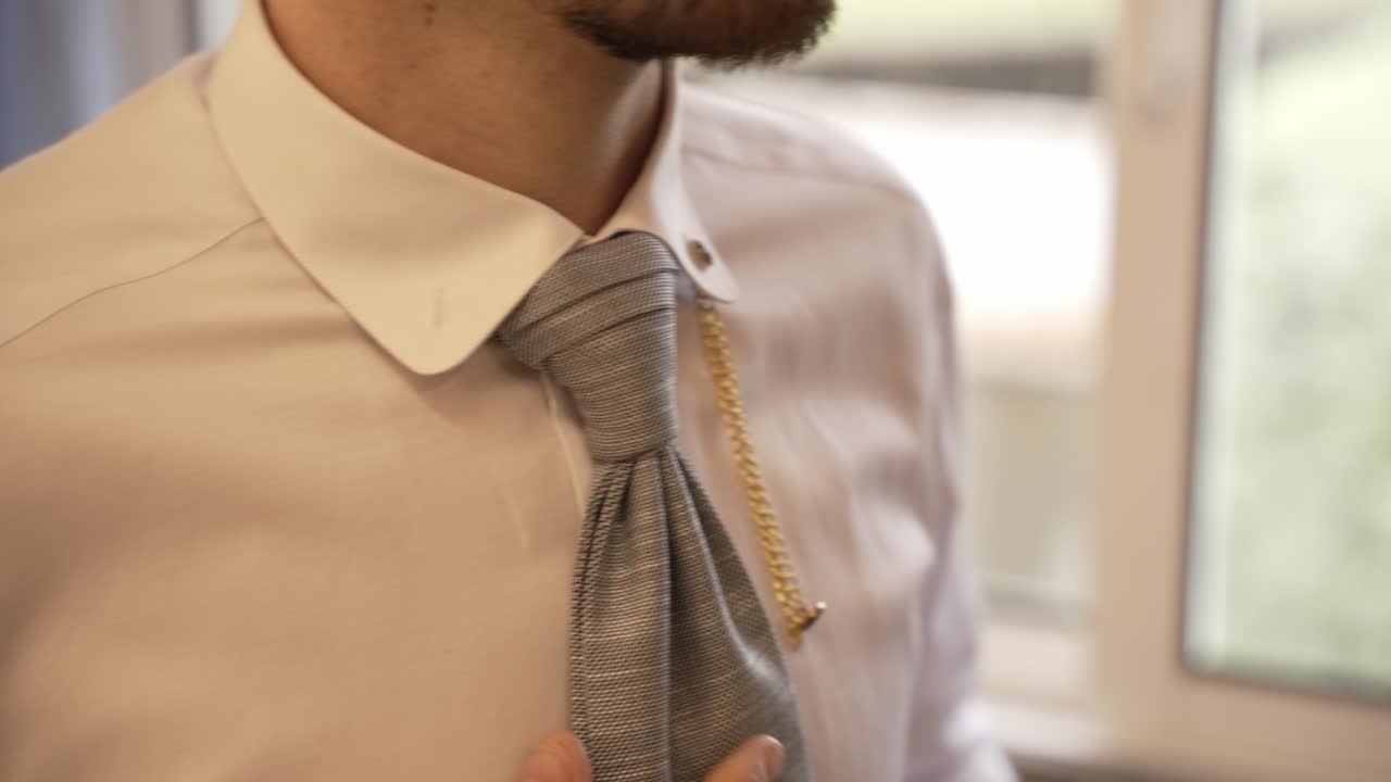 Close up of groom in white shirt adjusting gray tie with gold chain detail