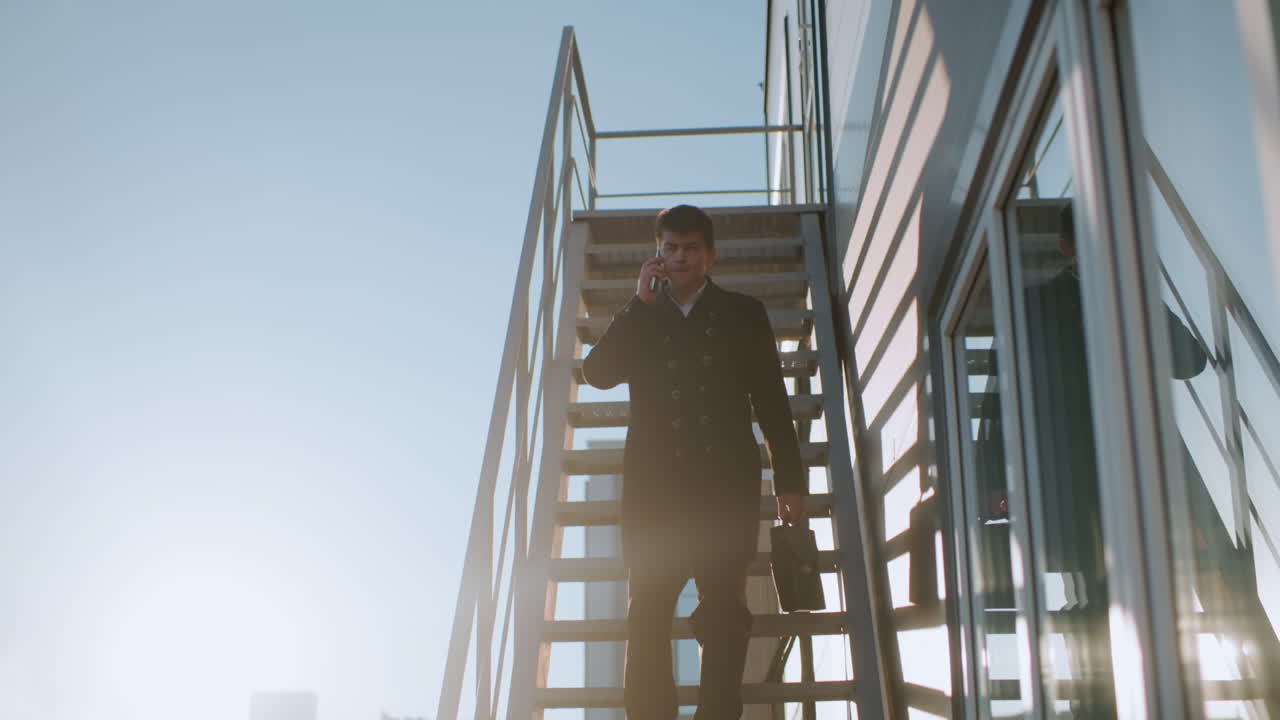 hombre con abrigo negro descendiendo la escalera en una llamada telefónica sosteniendo un maletín, asintiendo con la cabeza mientras habla bajo la luz del sol brillante, el fondo presenta vidrios reflectantes, coches estacionados y elementos urbanos