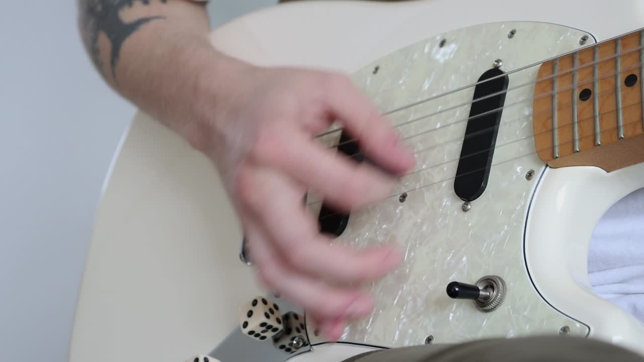 Una mano tiene un plettro e strimpella una chitarra elettrica, lo sfondo è neutro.