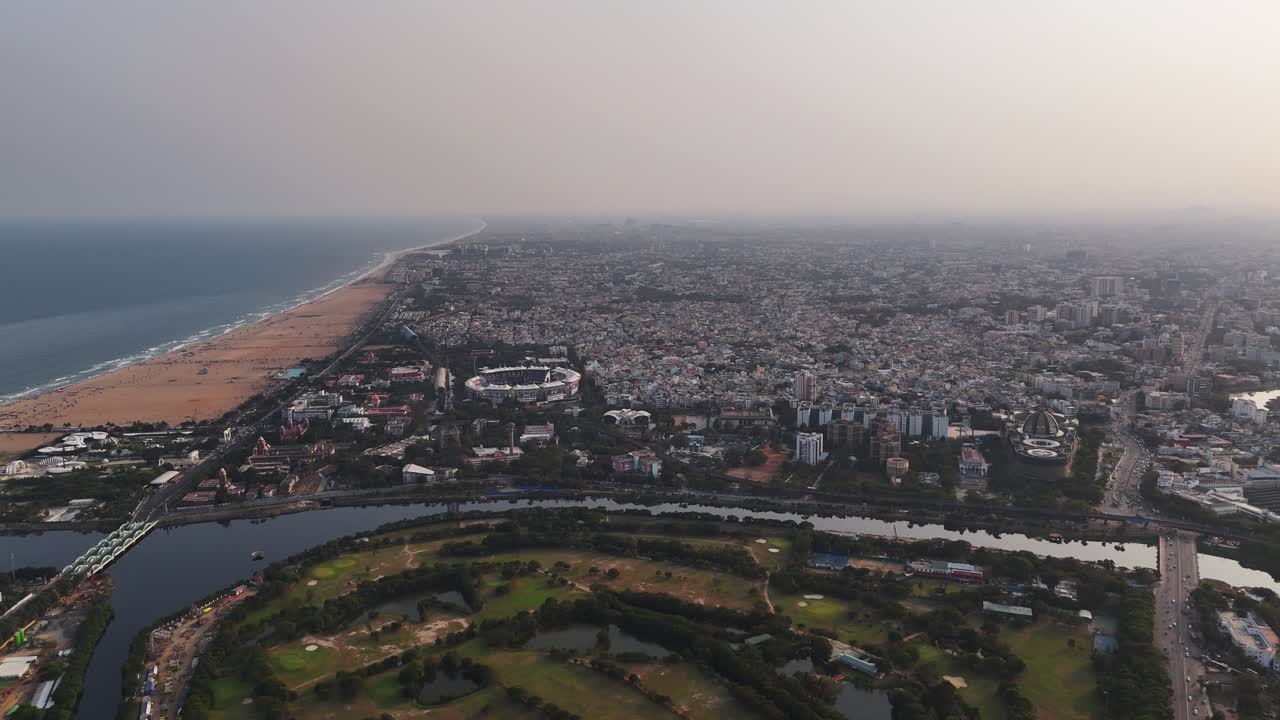 Una ripresa con drone sopra lo Stadio Chepauk da lunga distanza mostra l'intera arena di cricket con il suo tetto a baldacchino bianco e il campo centrale. La costa della Marina e gli edifici circostanti sono visibili oltre