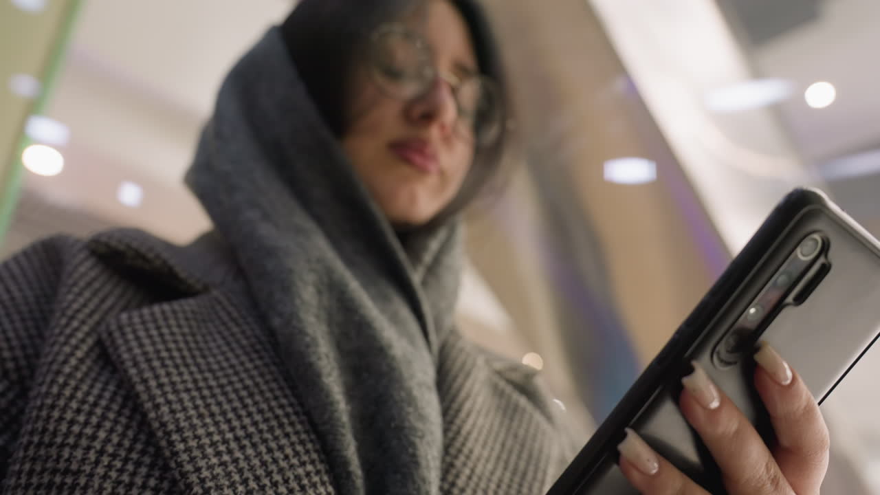 woman wearing scarf and checkered coat using smartphone while standing inside ascending elevator with soft focus lighting in background