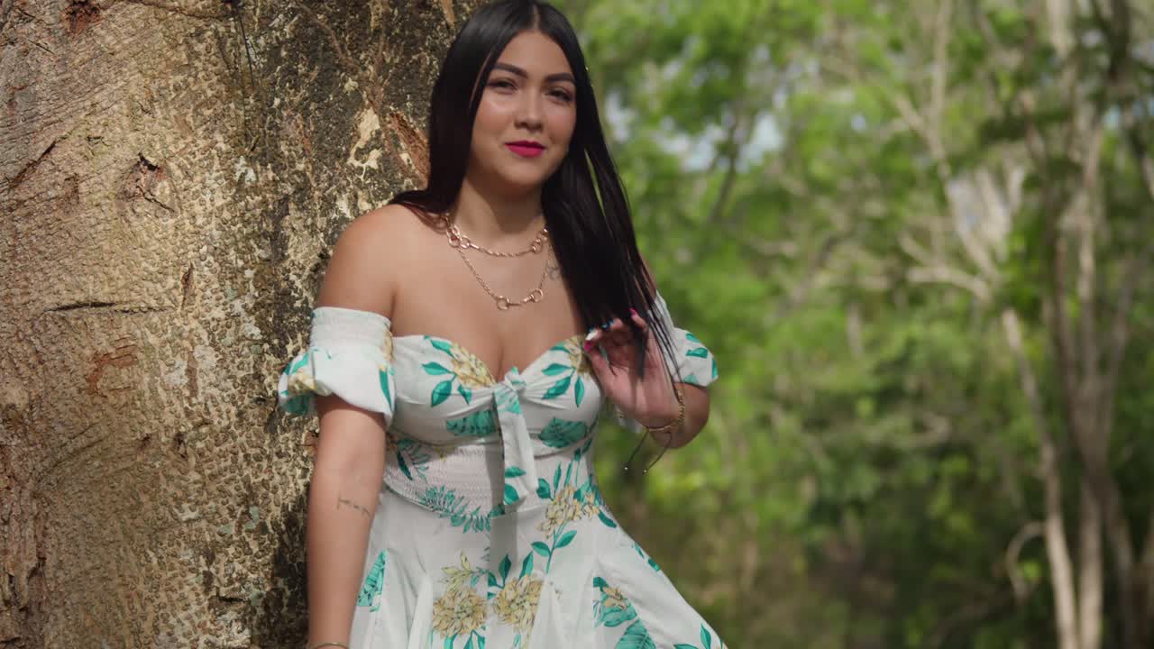 Jovem garota em um parque, usando um vestido curto, com um grande tronco de árvore ao fundo.