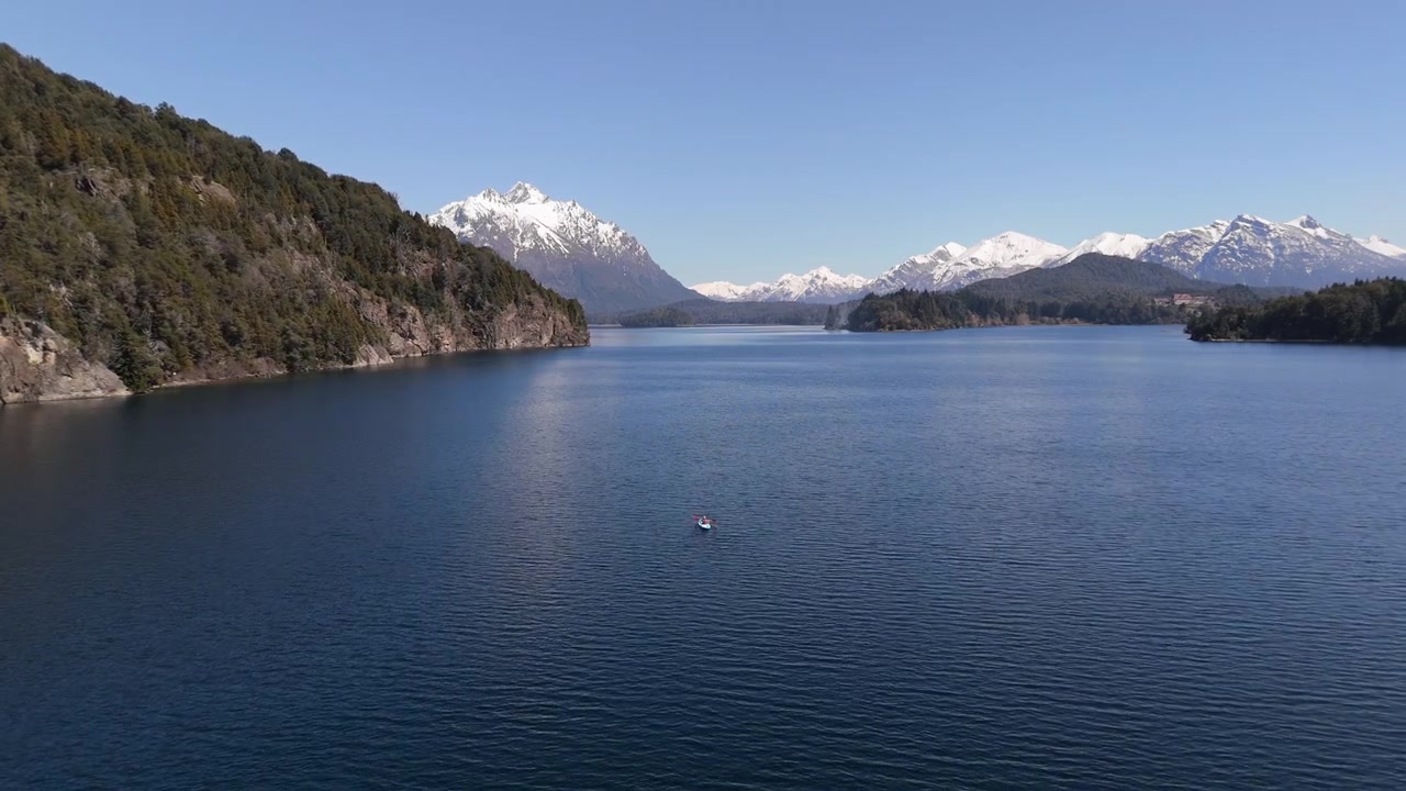 Vista aerea con zoom su un lago con uno sportivo o un uomo che fa kayak da solo, con paesaggio forestale e montagne innevate a Bariloche, Rio Negro, Argentina.