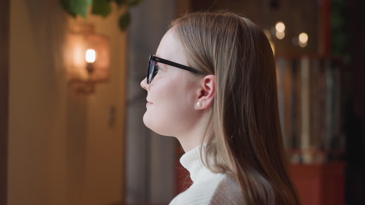 close up of smiling young woman in glasses turning gently toward camera with soft indoor lighting, blurred bokeh background, and cozy atmosphere creating warm relaxed mood