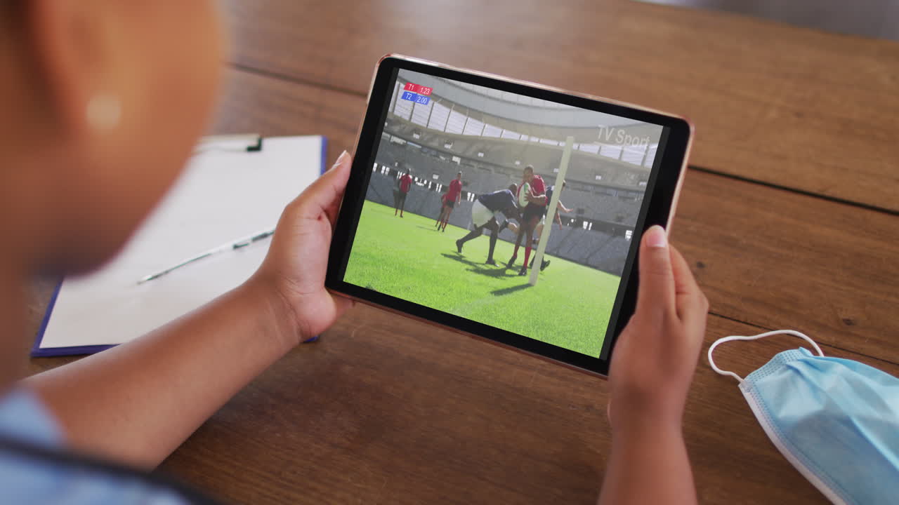 태블릿에서 럭비 경기를 시청하는 얼굴 마스크와 함께 테이블에 앉아있는 여성의 복합