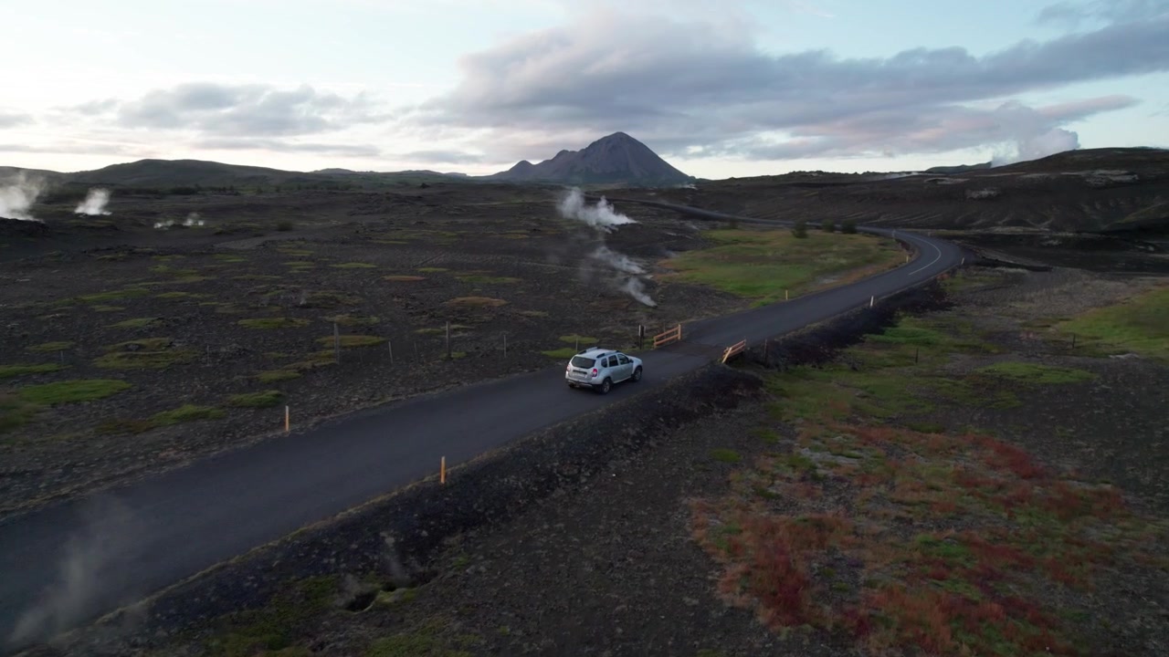 Ripresa aerea di inseguimento di un'auto che attraversa una remota area geotermica in Islanda, circondata da bocche fumanti e un aspro terreno vulcanico con vapore geotermico che si innalza tutt'intorno
