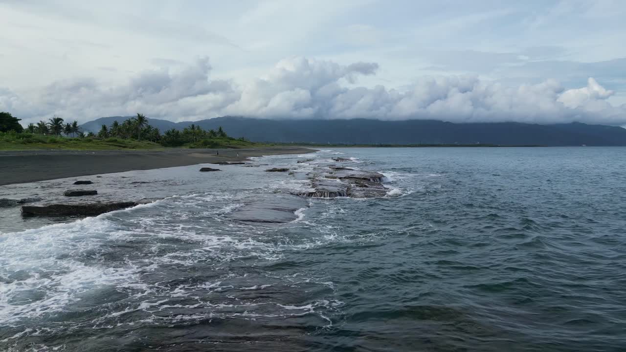 toma aérea de drones de olas que se estrellan contra las rocas de la costa en una playa idílica con montañas y cielos nublados en el fondo