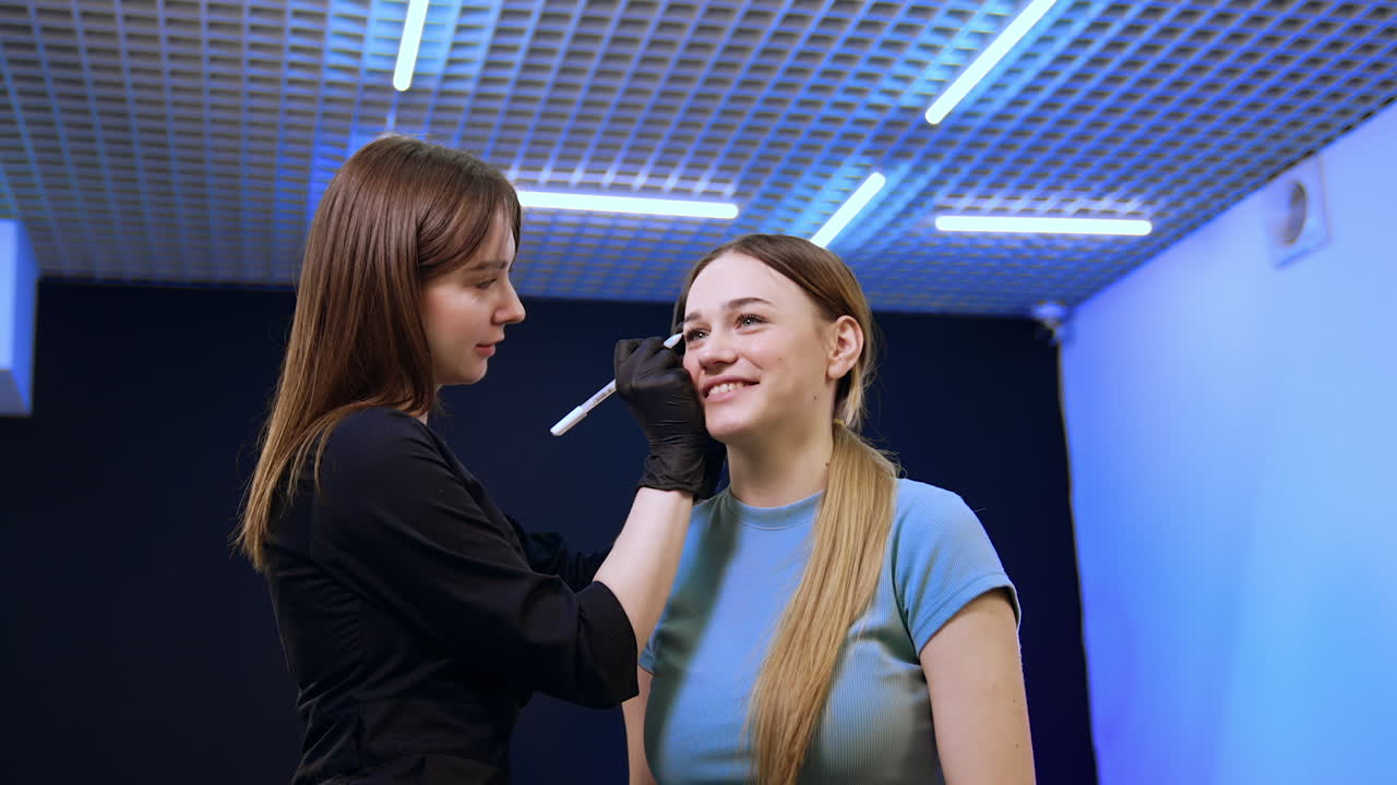 Long-haired brunette cosmetologist draws lines near the eyebrows of a client. Preparation for eyebrow permanent make-up. Low angle view.