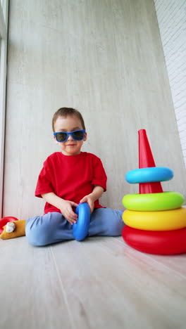 joyful caucasian kid wearing sunglasses rolls the pyramid rings by the floor. adorable baby playing at home. vertical video.
