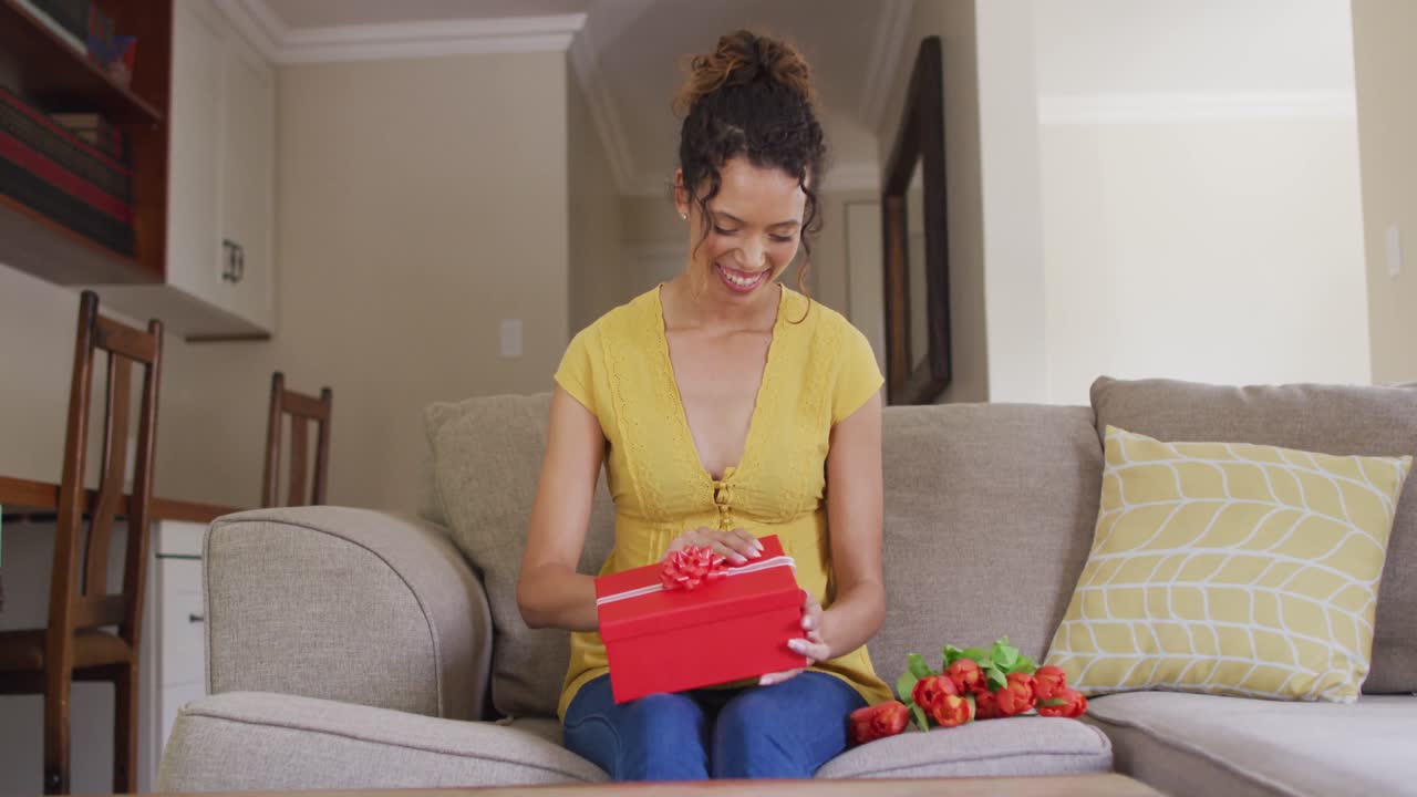 feliz mujer biracial sentada en el sofá, teniendo llamada de video de san valentín y apertura de regalo