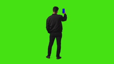 vista trasera de longitud completa del hombre sosteniendo el teléfono móvil de pantalla azul a la cámara contra el fondo de pantalla verde