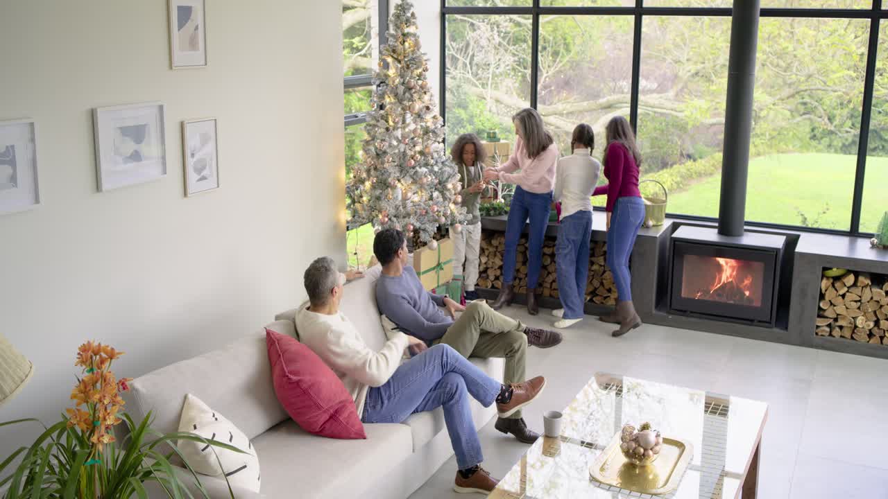 Diverse family at window placing gifts at Christmas tree, hanging ornaments father son sipping mugs