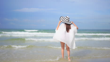 lonely woman on sandy beach running in fluttering tunic and floppy hat, raising hand full of joy in front of tropical sea waves, back view, selective focus, full frame