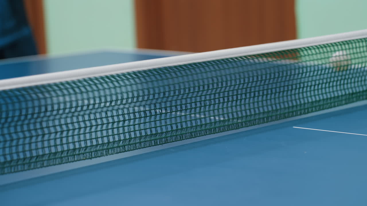 Close up of tennis ball dropping to ground after striking green net on blue table surface during indoor match capturing game detail, accuracy challenge