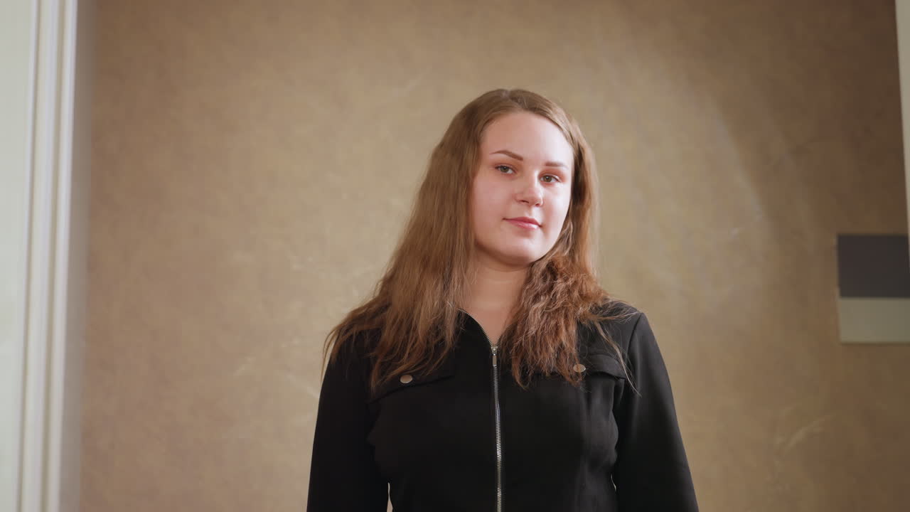 Zoom out portrait of beautiful model in black gown with front zipper, hand raised to adjust hair, posing with warm smile in soft indoor light, confident expression, elegant style, neutral background