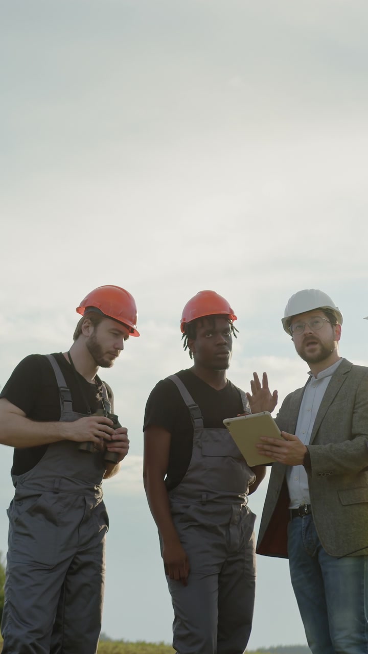 Diverse group of engineers and technicians in hard hats and overalls collaborating outdoors near