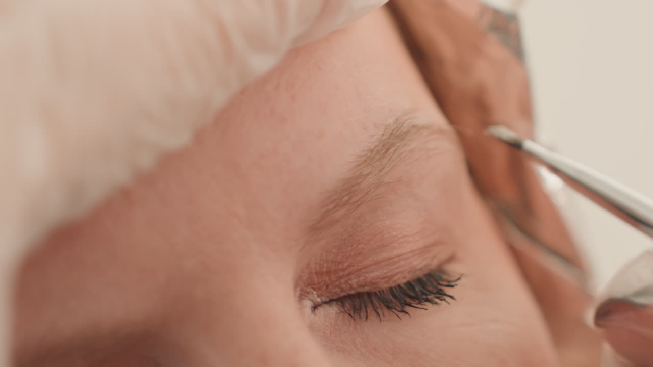 closeup eyebrow microblading on closed eye, esthetician applies pigment with fine tool wearing sterile gloves, soft neutral lighting, relaxed client, subtle contour enhancement, detailed precise