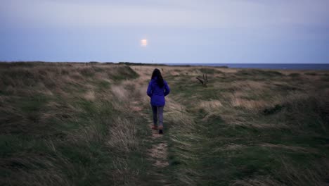 Anonymous-female-hiker-walking-on-dry-terrain-near-sea-in-evening