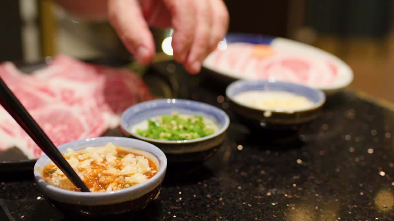 Hand adds garlic and herbs to dipping sauce beside raw meat under warm indoor lighting