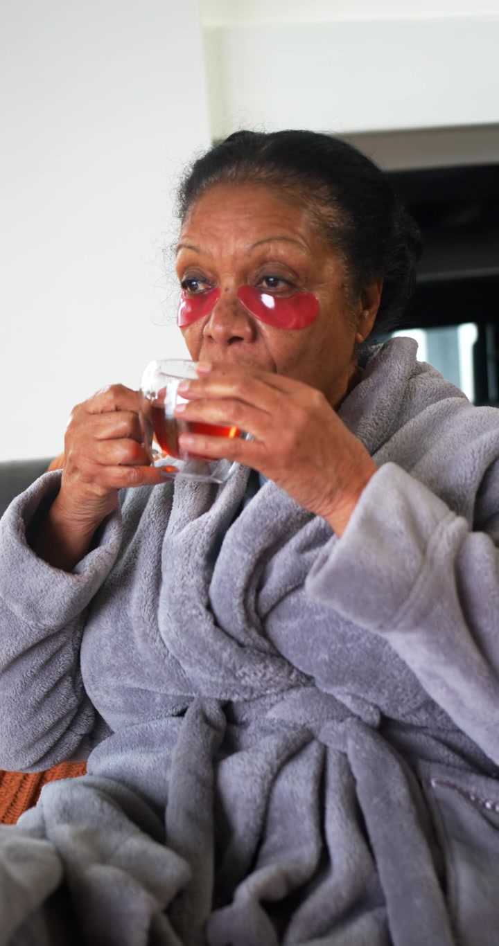 Vidéo verticale : Femme afro-américaine âgée en robe de chambre avec des coussins rouges, tenant une tasse, buvant à petites gorgées et se reposant sur un canapé
