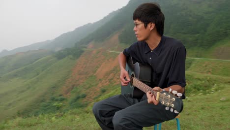 Video-Cinematográfico-De-Un-Joven-Coreano-Sentado-En-La-Cima-De-Un-Alto-Acantilado,-Tomando-Algo-De-Espacio-Mental-Y-Tocando-Su-Guitarra-Solo-Para-él-Mismo.