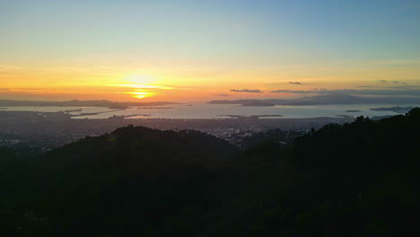 Drone-flying-backwards,-sunset-over-coastal-city-with-blue-skyline-in-the-background