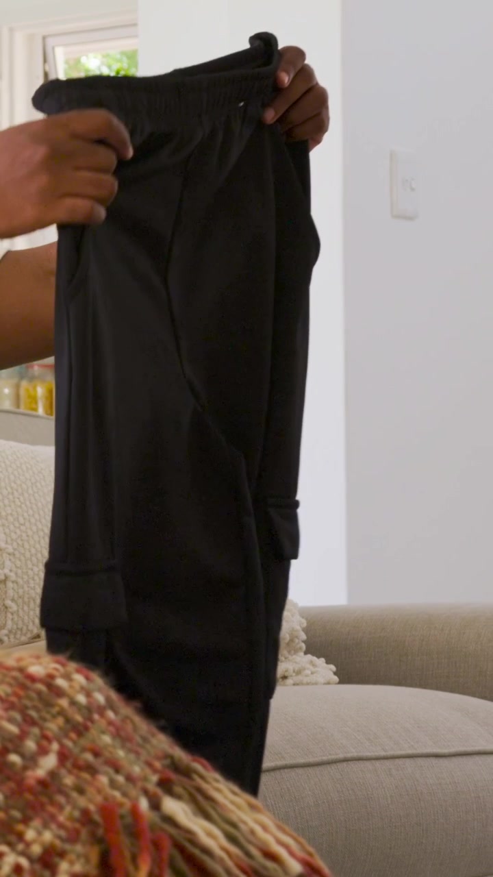 Vertical video: Pressing folded clothes, man in white polo glancing, unfolding dark pants on couch