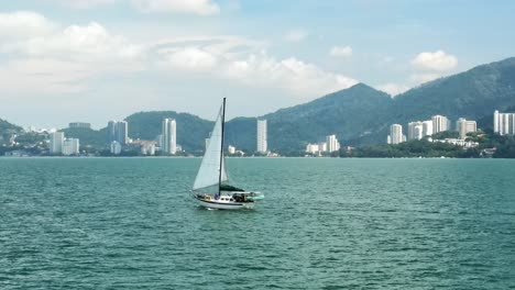 Sailboat-sails-across-the-calm-ocean-surface-with-Malaysia-City-Skyscrapers,-mountains,-and-tall-buildings-in-the-background