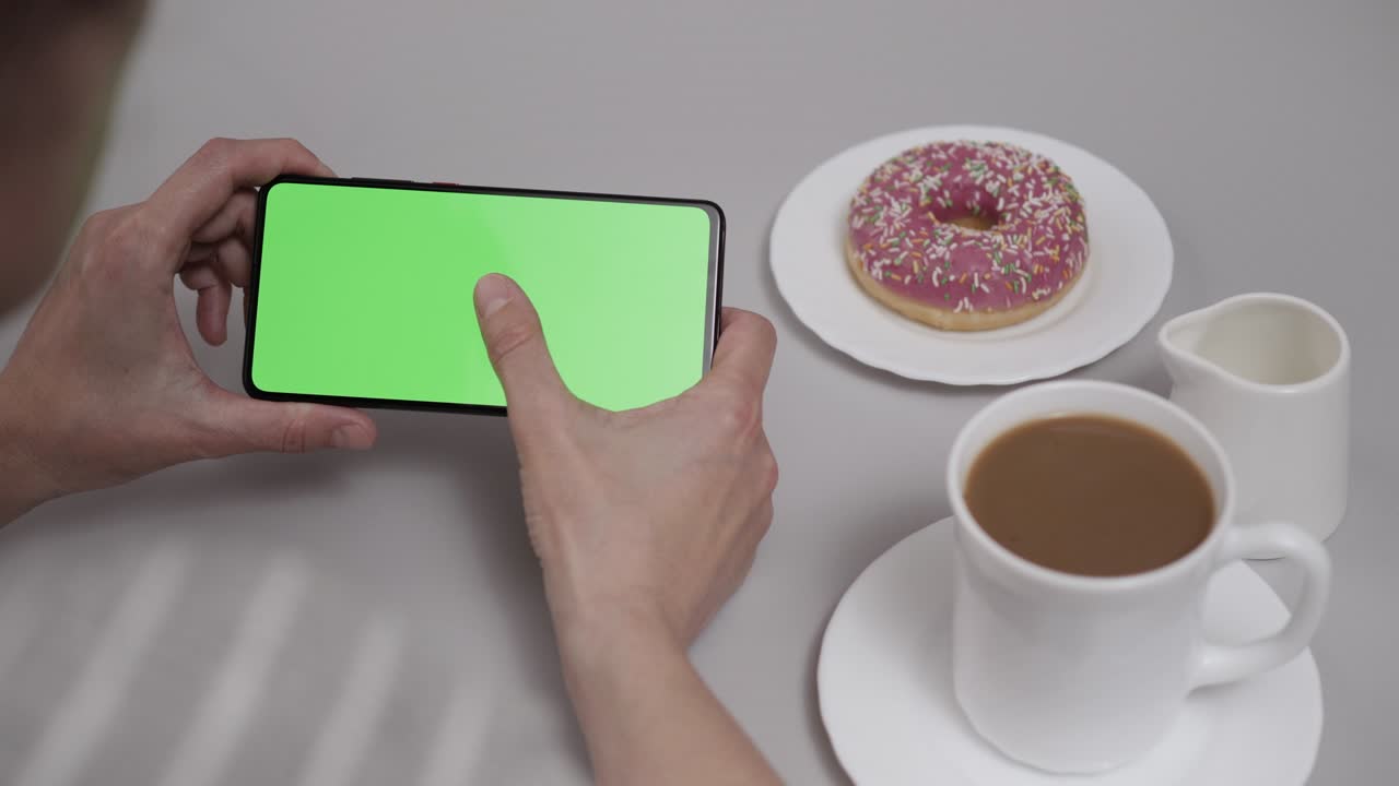 mujer sentada en la mesa de café donut usando un teléfono inteligente con pantalla verde de clave de croma, desplazándose a través de las redes sociales medios de comunicación en línea tienda internet. teléfono inteligente en modo horizontal con maqueta de pantalla verde.