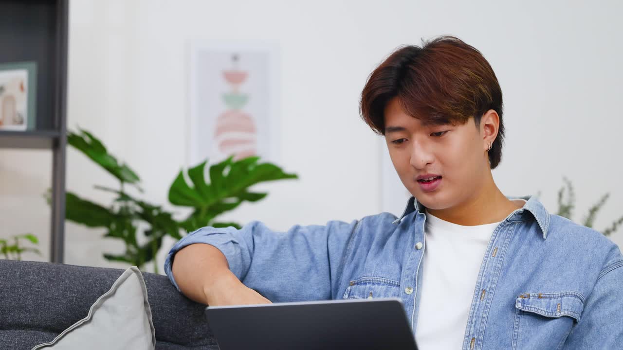 ソファに座り、ノートパソコンでのビデオ通話中に話したりジェスチャーしたりする若いアジア人男性