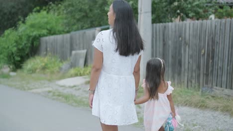 a pregnant woman and her young daughter, dressed in light summer outfits, walk hand-in-hand along a street in a residential neighborhood. the daughter holds a toy, enjoying the peaceful moment outdoor