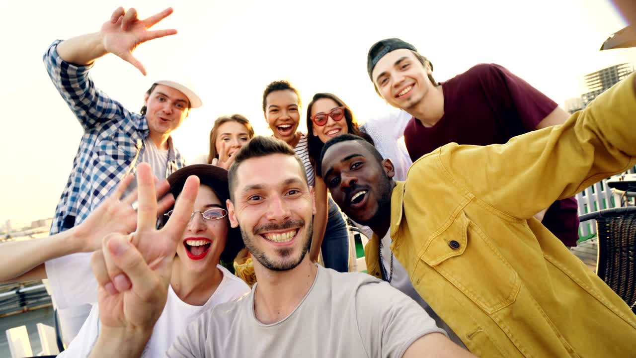 Point of view shot of bearded young man looking at camera, touching screen and adjusting camera then taking selfie with friends sitting on roof at table.