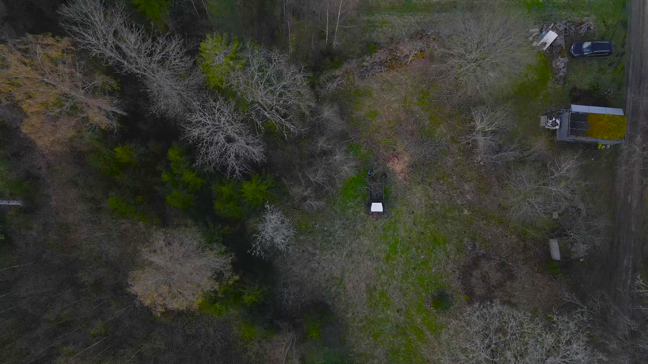 Aerial drone footage flying over a small retro and vintage tractor where people are throwing branches and garden waste, debris on during a cloudy sping day with leafless trees around them.