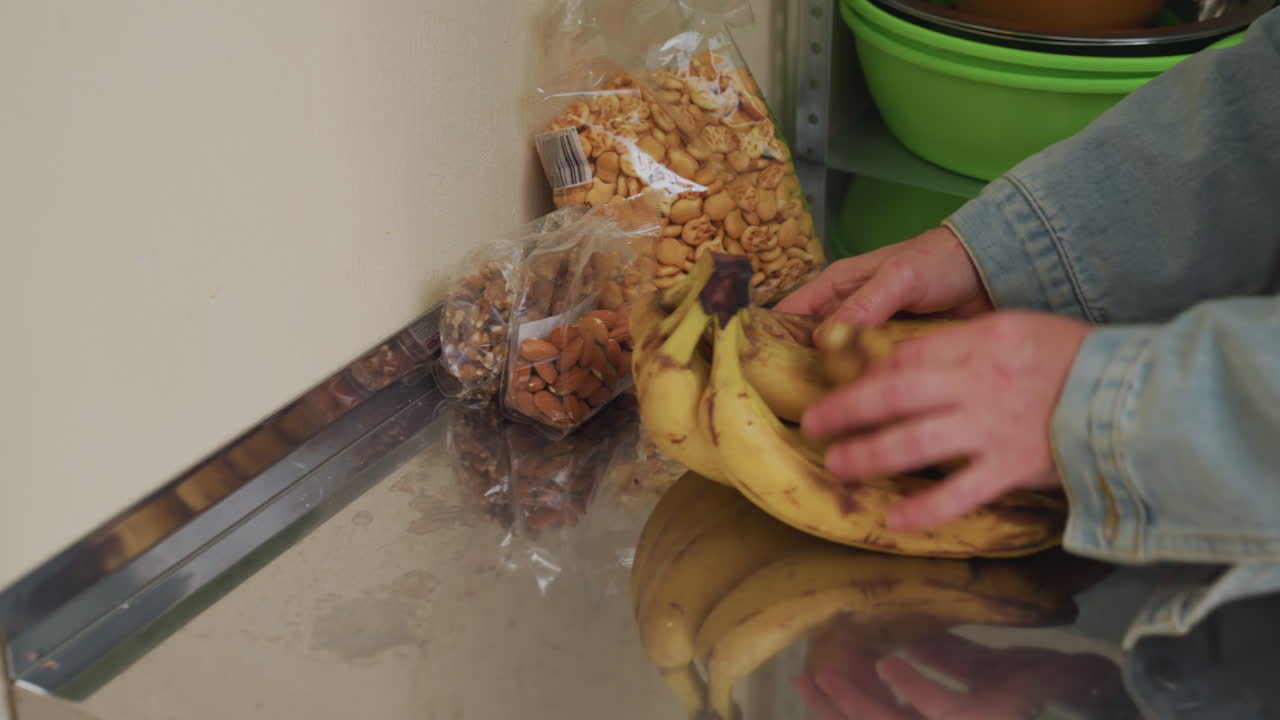hands place bananas then remove granola in compact storage area where denim jacket hands arrange produce and clear packaging, reflective metal countertop, green bowls on shelf, methodical restock